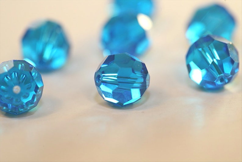 May include: A group of six turquoise blue faceted glass beads. The beads are round and have a shiny finish. They are scattered on a white surface.