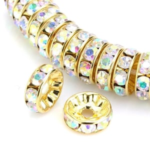May include: Gold-toned metal bead spacers with iridescent rhinestones. The beads are round with a flat top and a hole in the center. The rhinestones are arranged in a circle around the edge of the bead.