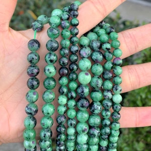 May include: A strand of green and black gemstone beads. The beads are round and have a speckled appearance.