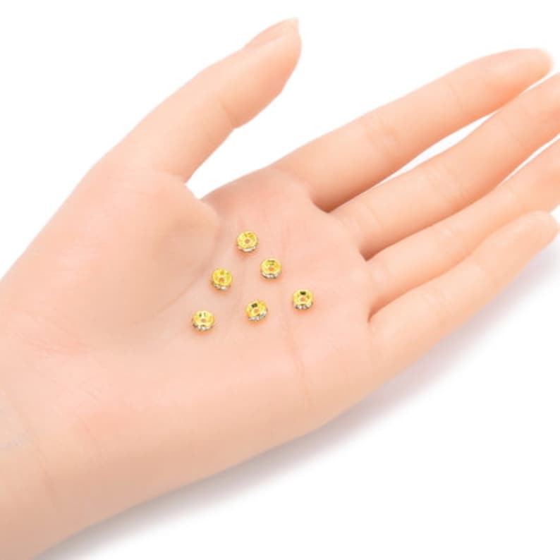 May include: A close-up of six gold-toned metal bead caps with clear rhinestones. The bead caps are scattered on a hand.