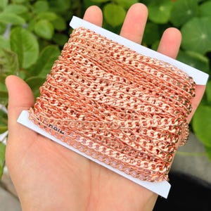 May include: A roll of rose gold curb chain, tightly wound on a white card. The chain links are uniform in size and shape, creating a continuous, textured surface. The chain is held in a hand, with a green leafy background.