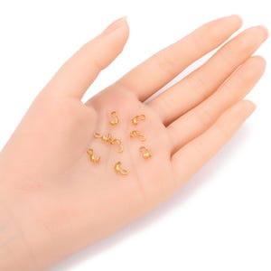 Könnte beinhalten: Nahaufnahme einer Hand, die eine Gruppe kleiner, goldfarbener Metallverschlüsse hält. Die Verschlüsse sind für die Schmuckherstellung oder andere Bastelprojekte gedacht.