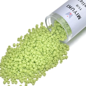 May include: A clear plastic container labeled "MIYUKI 11/0" is tipped over, spilling a pile of small, light green cylinder-shaped beads. The beads are scattered across a white surface, creating a textured pattern. The beads are for jewelry making.