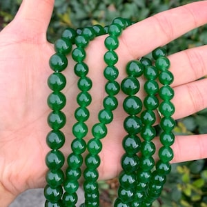 May include: A strand of green jade beads, strung on a cord and displayed on a hand. The beads are round and smooth, with a polished finish.
