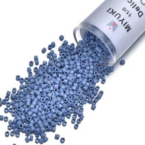 May include: A close-up shot shows a collection of small, light blue cylindrical beads spilling from a clear container. The container is labeled "MIYUKI 11/0 Delica". The beads are scattered on a white surface, suggesting their use in jewelry making or crafts.