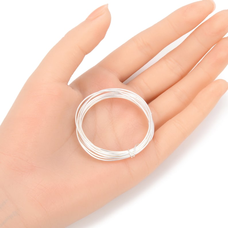 May include: A coil of silver-colored wire, tightly wound, rests in the palm of a hand. The wire appears to be thin and flexible, suitable for jewelry making or crafting. The bright metal contrasts with the skin tone.