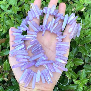 May include: A strand of iridescent purple amethyst crystal points, also known as amethyst aura quartz, strung on a clear thread. The points are various lengths and are arranged in a circular pattern.