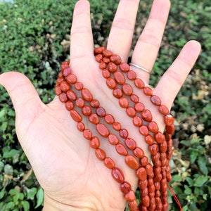 May include: A strand of red jasper beads, oval shaped, with a smooth, polished finish. The beads are strung on a thin cord and are displayed on a hand.