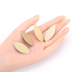 May include: Four gold-colored metal leaf-shaped charms with a hole at the top. The charms are on a hand.