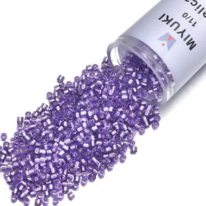 Peut inclure: Un récipient en plastique transparent de perles de rocaille Miyuki 11/0 se déverse sur une surface blanche. Les perles sont de couleur violet translucide. Le récipient porte une étiquette blanche avec le nom de la marque et la taille.