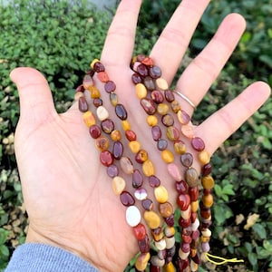 May include: A strand of multi-colored gemstone beads, featuring shades of red, orange, yellow, and brown. The beads are smooth and polished, with a natural, pebble-like shape.
