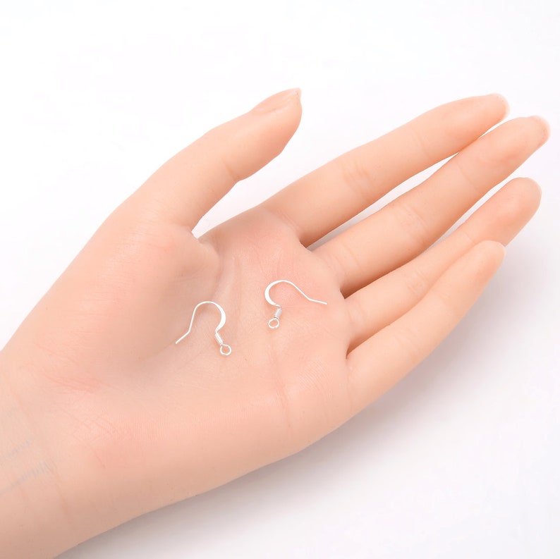 May include: Two silver earring hooks, also known as French hooks, are shown on a hand. The hooks are made of metal and have a small loop at the top.