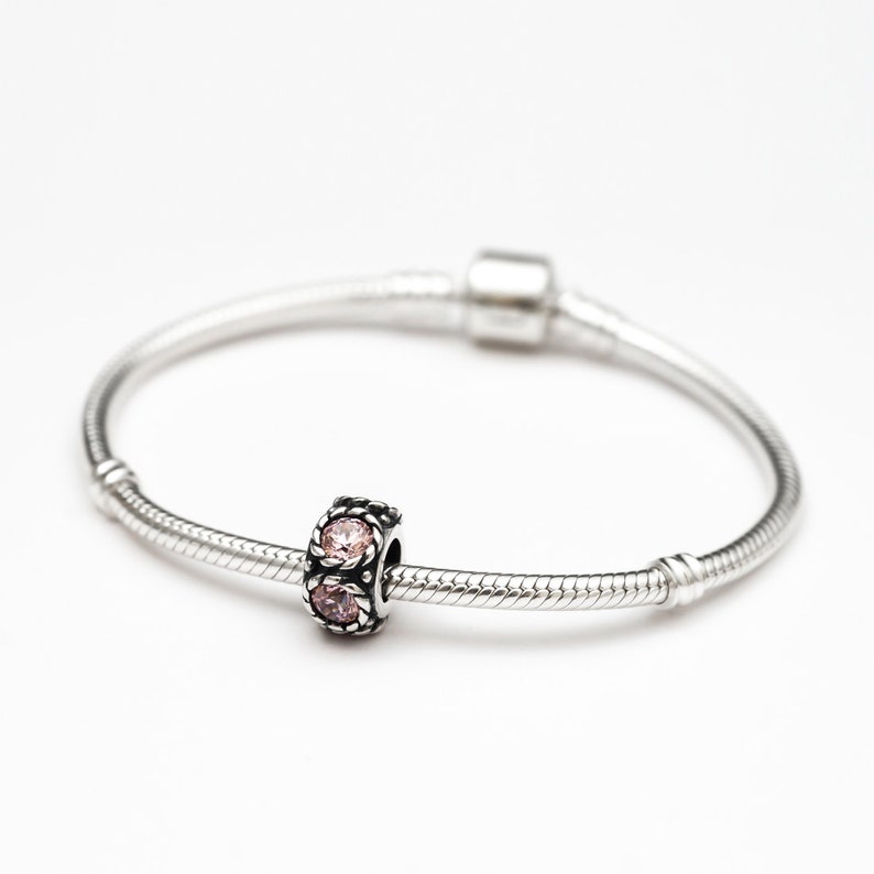 May include: A silver charm bracelet with a single charm featuring two pink gemstones set in a silver frame.