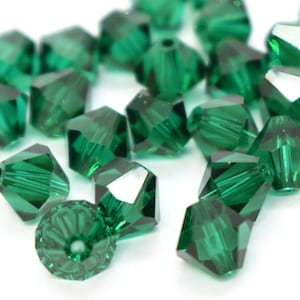 Puede incluir: Un grupo de cuentas de vidrio facetadas de color verde esmeralda. Las cuentas son pequeñas y tienen una forma puntiaguda. Están esparcidas sobre una superficie blanca.