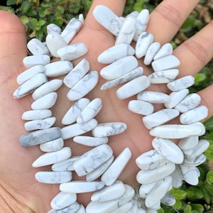 Peut inclure: Un brin de perles de pierre de howlite blanche, chaque perle est de forme ovale longue, irrégulière et légèrement pointue. Les perles ont une base blanche avec des veines grises dans toute leur longueur.