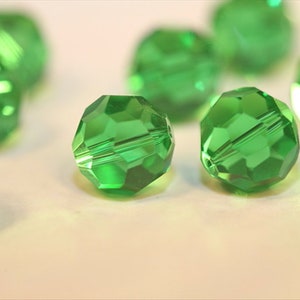 May include: A group of green faceted glass beads. The beads are round and have a shiny finish. The beads are scattered on a white surface.