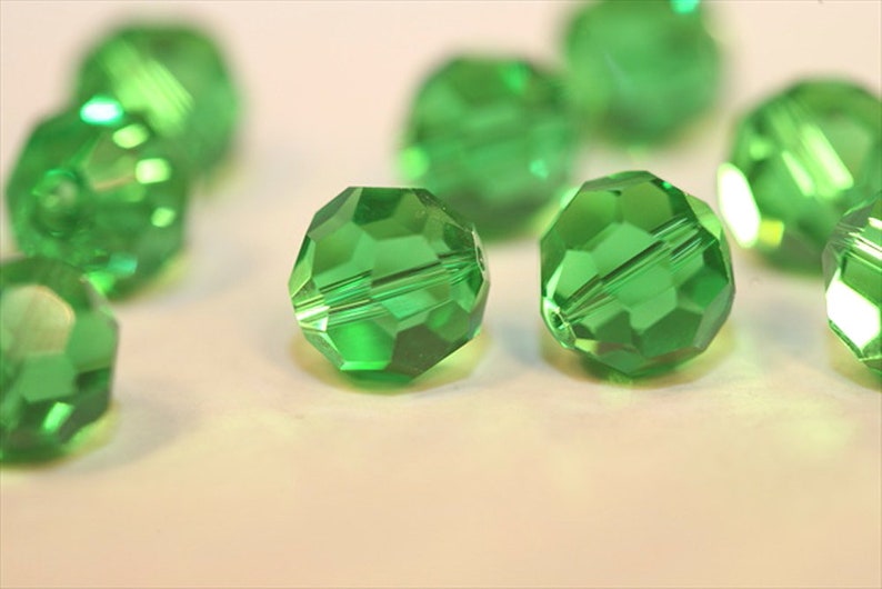 May include: A group of green faceted glass beads. The beads are round and have a shiny finish. The beads are scattered on a white surface.