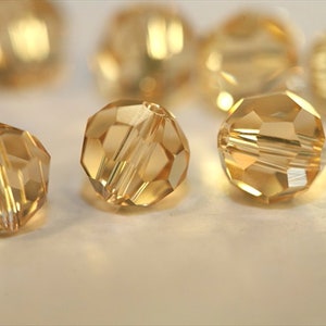 May include: A group of round, faceted, gold-toned glass beads. The beads are arranged on a white surface.