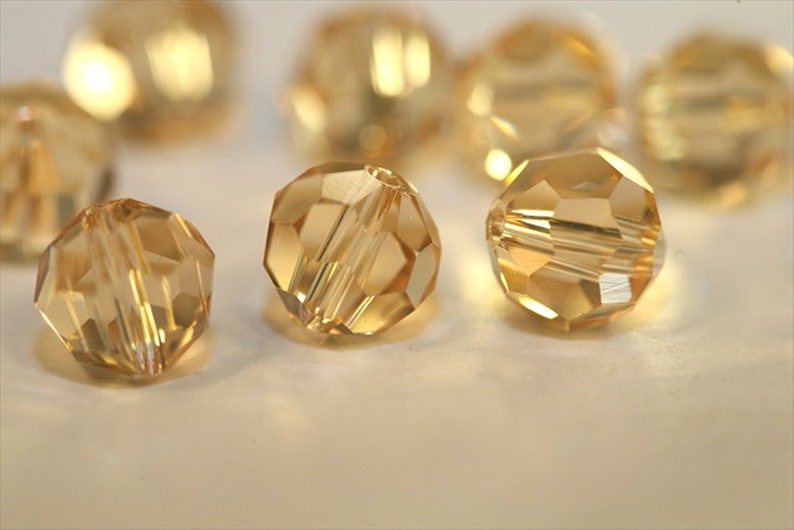 May include: A group of round, faceted, gold-toned glass beads. The beads are arranged on a white surface.