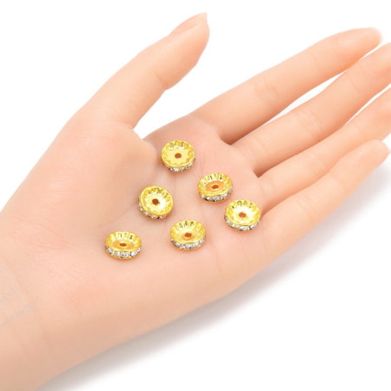 May include: A close-up of seven gold-toned metal bead caps with clear rhinestone accents. The bead caps are arranged on a hand.