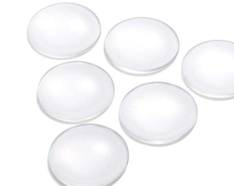 Pode incluir: Seis cabochões de vidro transparente, redondos e lisos, dispostos em semicírculo sobre um fundo branco.