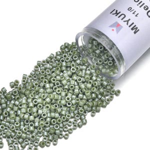 May include: A clear plastic container of olive green Miyuki Delica beads, size 11/0, spilling onto a white surface. The beads are cylindrical with a metallic sheen. The container has a white label with the brand name and product details.