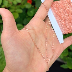 Könnte beinhalten: Roségoldfarbene Kette mit kleinen, ovalen Gliedern. Die Kette wird in einer Hand gehalten, mit einer Rolle passender Kette im Hintergrund. Die Kette ist filigran und scheint neu zu sein.