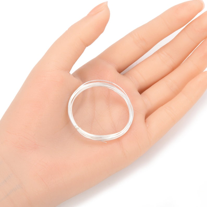 May include: A coil of thin, silver-colored wire rests in an open hand. The wire is tightly wound, forming a circular shape. The background is a plain, light color, highlighting the wire's metallic sheen. This wire is likely used for jewelry making or crafts.