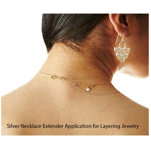 May include: A close-up of a person's neck wearing three necklaces, including a silver necklace extender. The extender is a delicate chain with a small pearl charm. The person is also wearing a pair of silver earrings.
