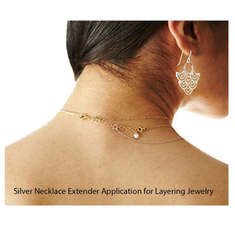 May include: A close-up of a person's neck wearing three necklaces, including a silver necklace extender. The extender is a delicate chain with a small pearl charm. The person is also wearing a pair of silver earrings.