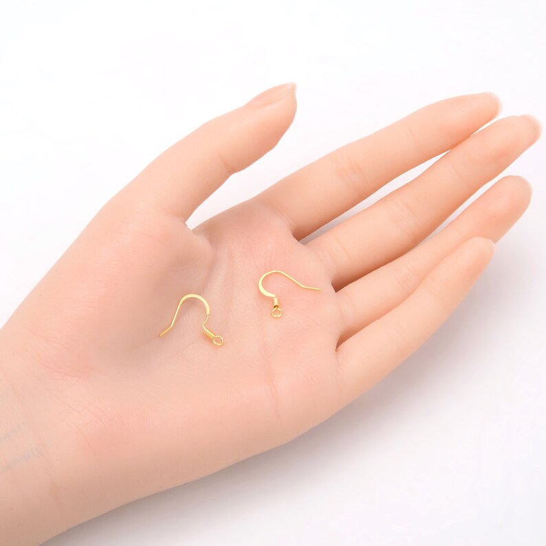 May include: Two gold-toned earring hooks, one with a loop at the end and one with a straight end, are shown on a hand.