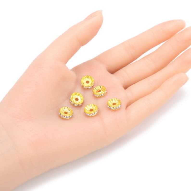 May include: A close-up of six gold-toned metal bead caps with clear rhinestones. The bead caps are round and have a flat top and bottom.