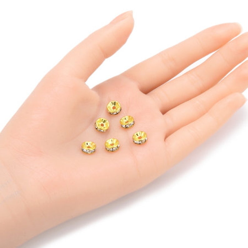 May include: Six gold-toned metal bead caps with clear rhinestone accents. The bead caps are arranged in a semi-circle on a hand.