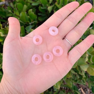 May include: Six pink donut-shaped beads made of rose quartz. The beads are smooth and polished.