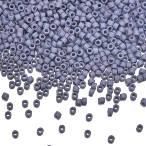 May include: A close-up shot of a pile of small, cylindrical beads in a muted blue-gray color. The beads are uniform in size and shape, with a hole running through the center. They are scattered across the surface.