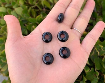 May include: Five black donut-shaped beads with a hole in the center. The beads are on a person's hand.