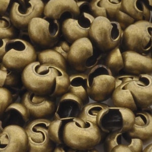 May include: A close-up of a pile of antique brass crimp beads. The beads are small and round with a small opening on one side. They are perfect for adding a touch of vintage style to jewelry making projects.