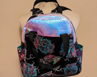 May include: A black backpack with a holographic iridescent front pocket and a colorful print of a band on the main compartment. The print features a lightning bolt design and the word "Queen".
