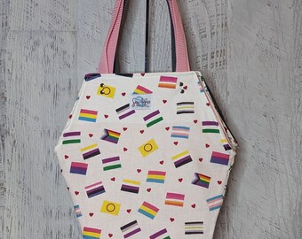 May include: A white tote bag with a coffin shape. The bag is decorated with a variety of pride flags and small red hearts. The bag has pink and black straps.