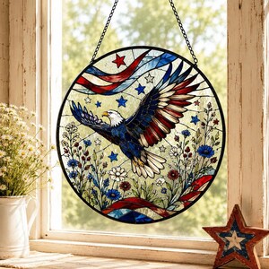 Könnte beinhalten: Ein runder Buntglas-Sonnenfänger mit einem fliegenden Weißkopfseeadler mit ausgebreiteten Flügeln. Das Design umfasst rote, weiße und blaue Farben, Sterne und florale Elemente. Das Stück ist in schwarzes Metall gefasst und hängt an einer schwarzen Kette.