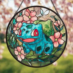 Może przedstawiać: Okrągłe witrażowe okno przedstawiające turkusową postać Bulbasaur otoczoną różowymi kwiatami i zielonymi liśćmi. Dzieło sztuki jest oprawione w czarną metalową ramę i zawieszone na łańcuchu. Tło to miękkie rozmycie różu i zieleni.