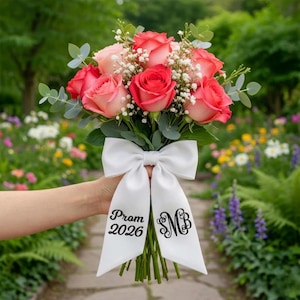 May include: A bouquet of red and pink roses, baby's breath, and greenery. The bouquet is tied with a white satin bow with the text "Prom 2026" and the initials "MB" in black script. The flowers are held in front of a blurred garden background.