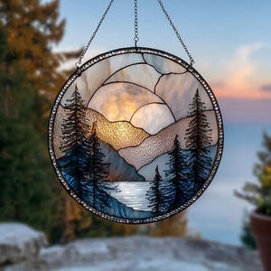 Könnte beinhalten: Rundes Buntglasfenster mit einer Berglandschaft bei Sonnenuntergang. Das Design zeigt silhouettierte Kiefern, einen See und geschichtete Berge in Blau-, Weiß- und Bernsteintönen. Das Fenster hat einen schwarzen Metallrahmen und eine Kette zum Aufhängen.