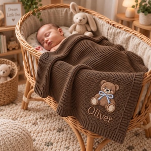 May include: A dark brown knitted baby blanket featuring a textured waffle pattern, a light brown teddy bear appliqué with a blue ribbon, and the name Oliver embroidered in a light brown script font, draped over a wicker bassinet.