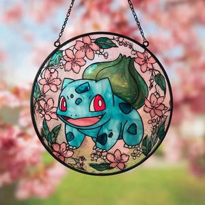 Może przedstawiać: Okrągłe okno witrażowe z turkusową postacią Bulbasaur, otoczone różowymi kwiatami wiśni i zielonymi liśćmi. Okno jest oprawione w czarną ramę i zawieszone na łańcuchu.
