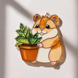 以下が含まれることがあります： 植木鉢を持ったハムスターの形をした装飾ピン。 ハムスターは茶色、タン、白の毛皮、黒い目、ピンクの足を持っています。 植物は緑色で、鉢は茶色です。 ピンは金色の輪郭です。