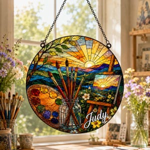 May include: A circular stained glass suncatcher featuring a landscape painting on an easel, a glass jar filled with paintbrushes, and a paint palette. The design includes a sunrise over mountains and the name Judy in white script lettering.