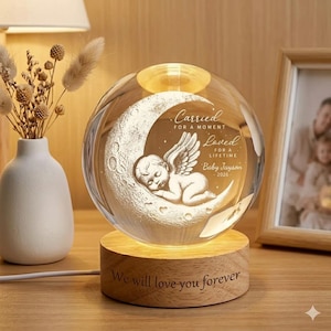 May include: A crystal sphere featuring a sleeping angel on a crescent moon, lit from within. The sphere is engraved with the words "Carried for a moment, Loved for a lifetime, Baby Jayson 2026." It rests on a wooden base inscribed with "We will love you forever."