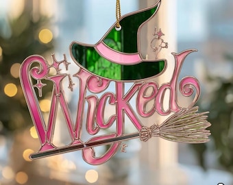 Puede incluir: Adorno decorativo con la palabra "Wicked" en escritura rosa, un sombrero de bruja verde y rosa y una escoba dorada. El adorno cuelga de una cadena dorada. Adecuado para Halloween.