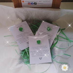 Könnte beinhalten: Drei kleine weiße Karten mit dem Text "Good Luck" und einem grünen Edelstein. Jede Karte enthält eine Botschaft über Selbstvertrauen und Unterstützung. Die Karten sind in durchsichtigem Kunststoff verpackt und von grünen Papierschnipseln umgeben.
